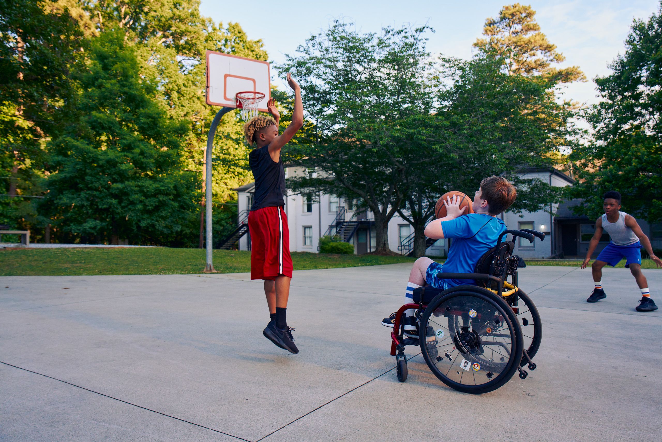 Three teens play basketball at a neighborhood park. One teen, a white boy seated in a manual wheelchair, holds the ball, ready to shoot. A second teen, a light-skinned African American male, jumps forward with arms outstretched, ready to defend the shot. A third teen, a Black boy, crouches, ready to receive a pass. The teen seated in a manual wheelchair has short brown hair and is wearing blue athletic wear. The second teen defending has curly brown to blonde hair is wearing black and red athletic wear. The third teen has short natural black hair styled high on top and is wearing gray and blue athletic wear.