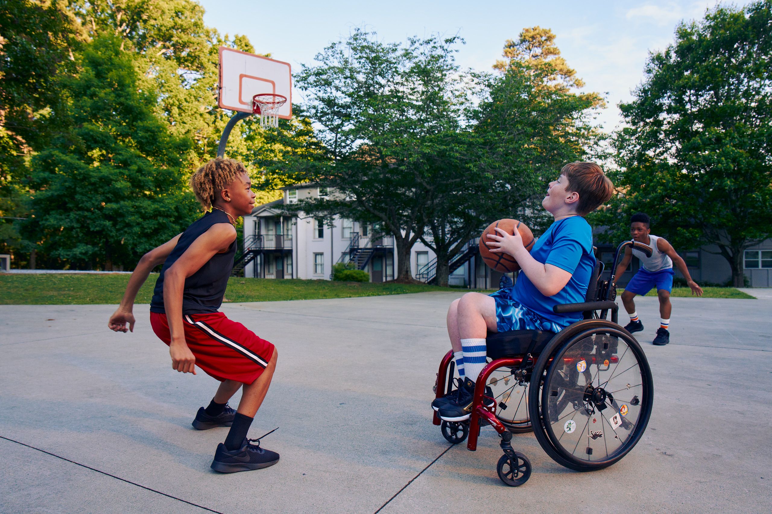 Three teens play basketball at a neighborhood park. One teen, a white boy seated in a manual wheelchair, holds the ball, ready to shoot. A second teen, a light-skinned African American male, crouches in a jumping position. A third teen, a Black boy, crouches, ready to receive a pass. The teen seated in a manual wheelchair has short brown hair and is wearing blue athletic wear. The second teen ready to jump has curly brown to blonde hair is wearing black and red athletic wear. The third teen has short natural black hair styled high on top and is wearing gray and blue athletic wear.
