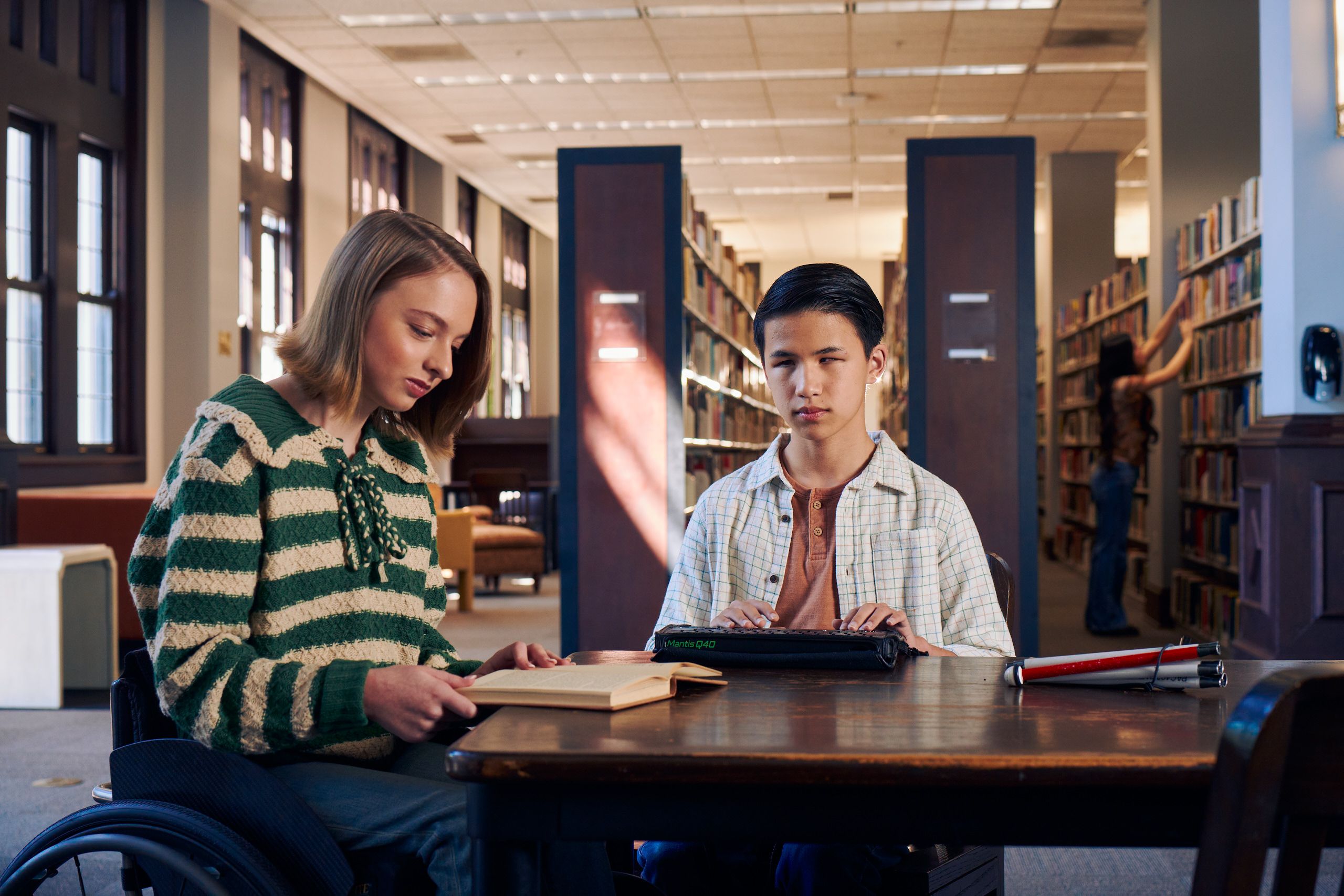 A white teenage girl seated in a manual wheelchair reads a book with focus at a table in a library. Next to her, a Chinese teen, who is a blind cane user, reads on a refreshable braille display. His white cane is collapsed and is placed the table next to him. The teen girl seated in a manual wheelchair has a dark blonde bob and is wearing a green and white striped knit shirt and jeans. The teen boy has slicked back black hair and a friendly smile with braces. He is wearing a rust colored shirt under a white plaid button down shirt.
