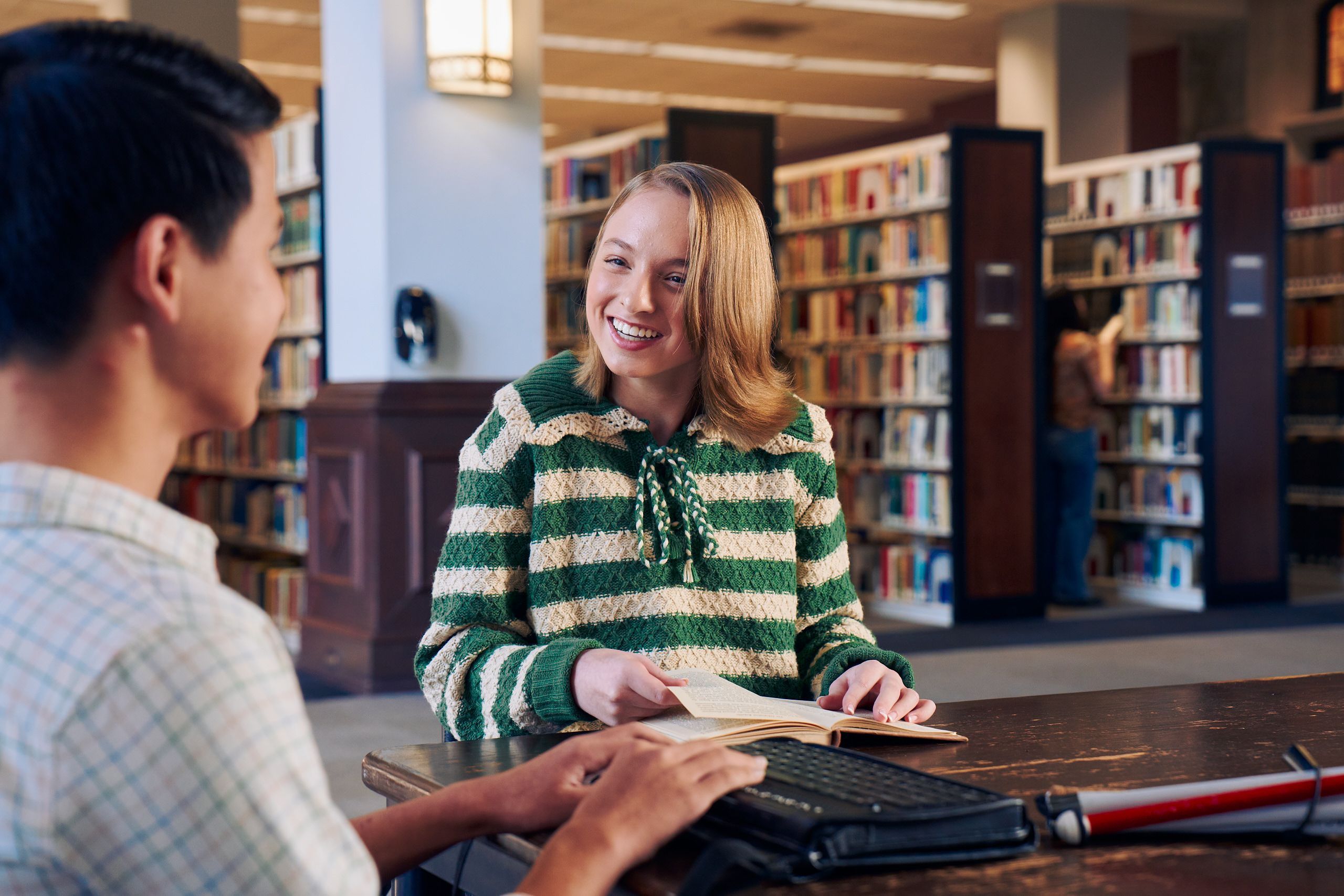 A white teenage girl seated in a manual wheelchair smiles brightly at a friend and holds a book open at a table in a library. Her friend, a Chinese teen, who is a blind cane user, has a refreshable braille display in front of him. His white cane is collapsed and is placed the table next to him. The teen girl has a dark blonde bob and is wearing a green and white striped knit shirt and jeans. The teen boy has slicked back black hair and is wearing a white plaid button down shirt.