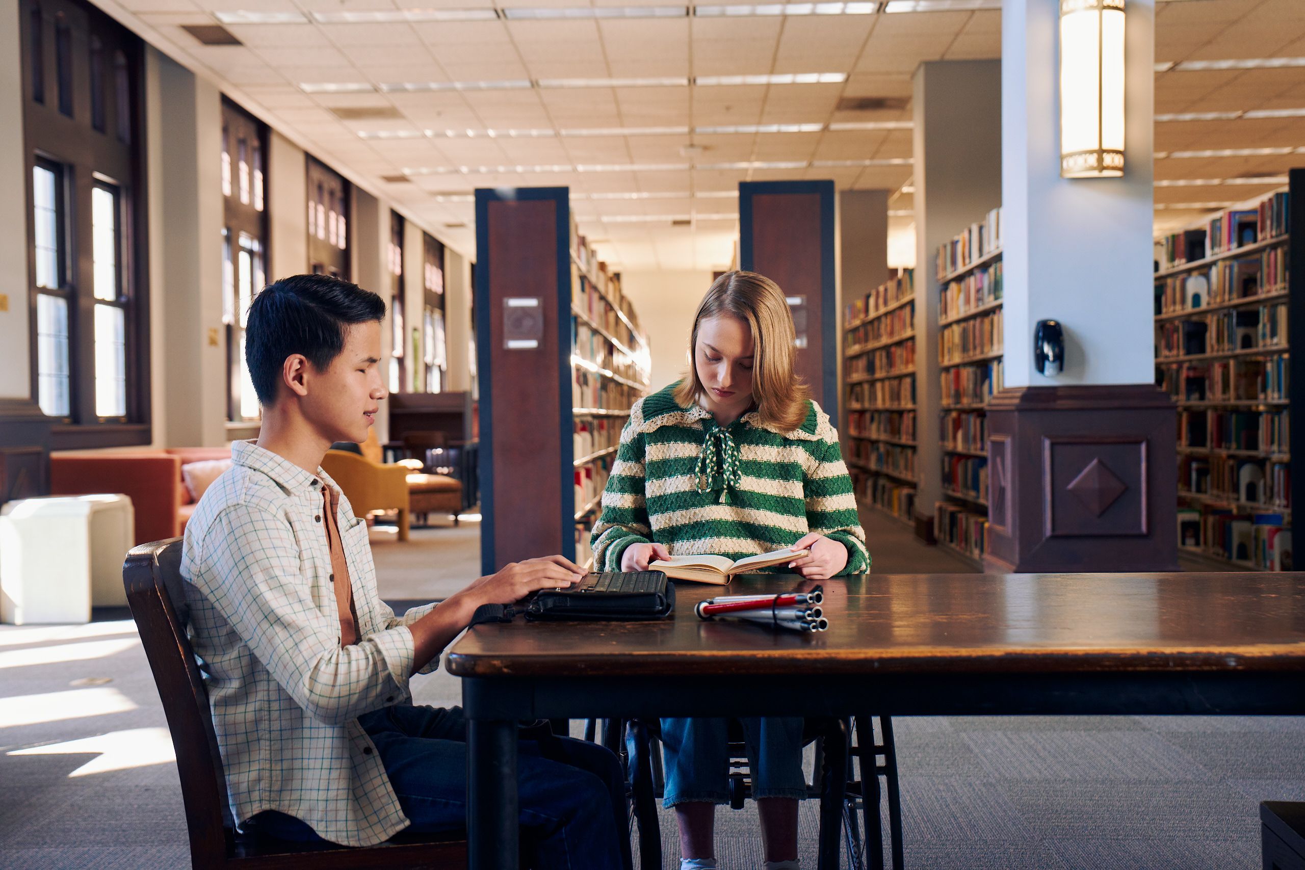 Two teenage friends read at a table in a library. One teen, a Chinese teen, who is a blind cane user, reads on a refreshable braille display. His white cane is collapsed and is placed the table next to him. The other teen, a white teenage girl seated in a manual wheelchair, is seated in a manual wheelchair and holds a hardcover book open. The teen girl has a dark blonde bob and is wearing a green and white striped knit shirt and jeans. The teen boy has slicked back black hair and is wearing a white plaid button down shirt.