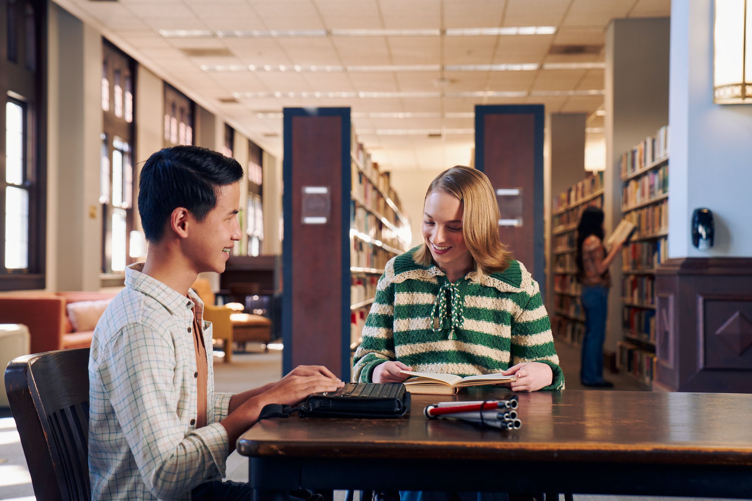 Two teenage friends laugh and read at a table in a library. One teen, a Chinese teen, who is a blind cane user, reads on a refreshable braille display. His white cane is collapsed and is placed the table next to him. The other teen, a white teenage girl seated in a manual wheelchair, is seated in a manual wheelchair and looks down at a hardcover book she has open. The teen girl has a dark blonde bob and is wearing a green and white striped knit shirt and jeans. The teen boy has slicked back black hair and is wearing a white plaid button down shirt.