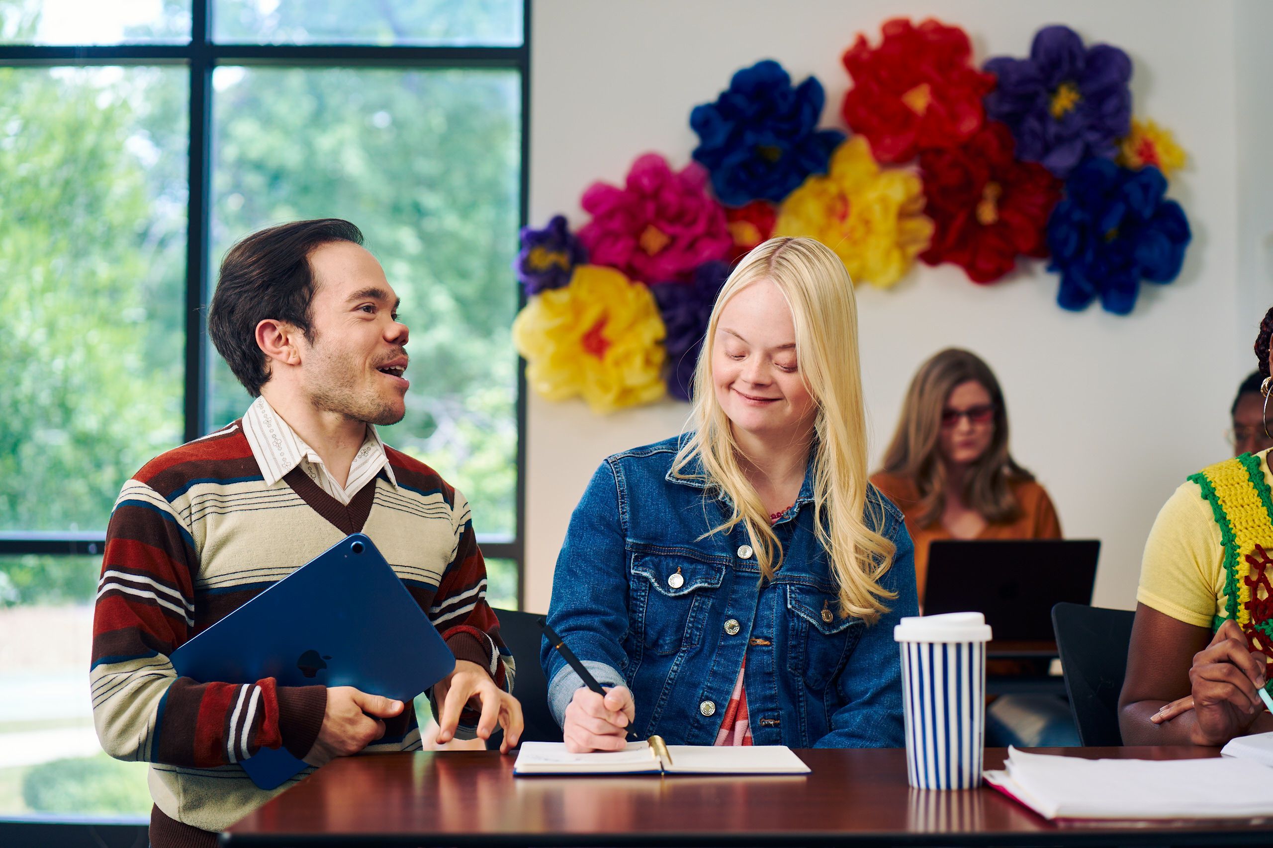 A caucasian college student with Down Syndrome sits at a lecture table and takes notes while her professor, who is standing next to her, chats with her. She has long blonde hair, blue eyes, and is wearing a jean hacket and several bright scrunchies on her wrist. Another student behind her, a blind white woman wearing glasses with red lenses, types on a laptop. A large flower sculpture hangs on the wall behind the desks. Her professor, a white man with medium brown hair and light brown eyes who has dwarfism, smiles and holds a tablet in his hand. He has short brown hair and is wearing a red striped sweater, a collared shirt, and dark dress pants.