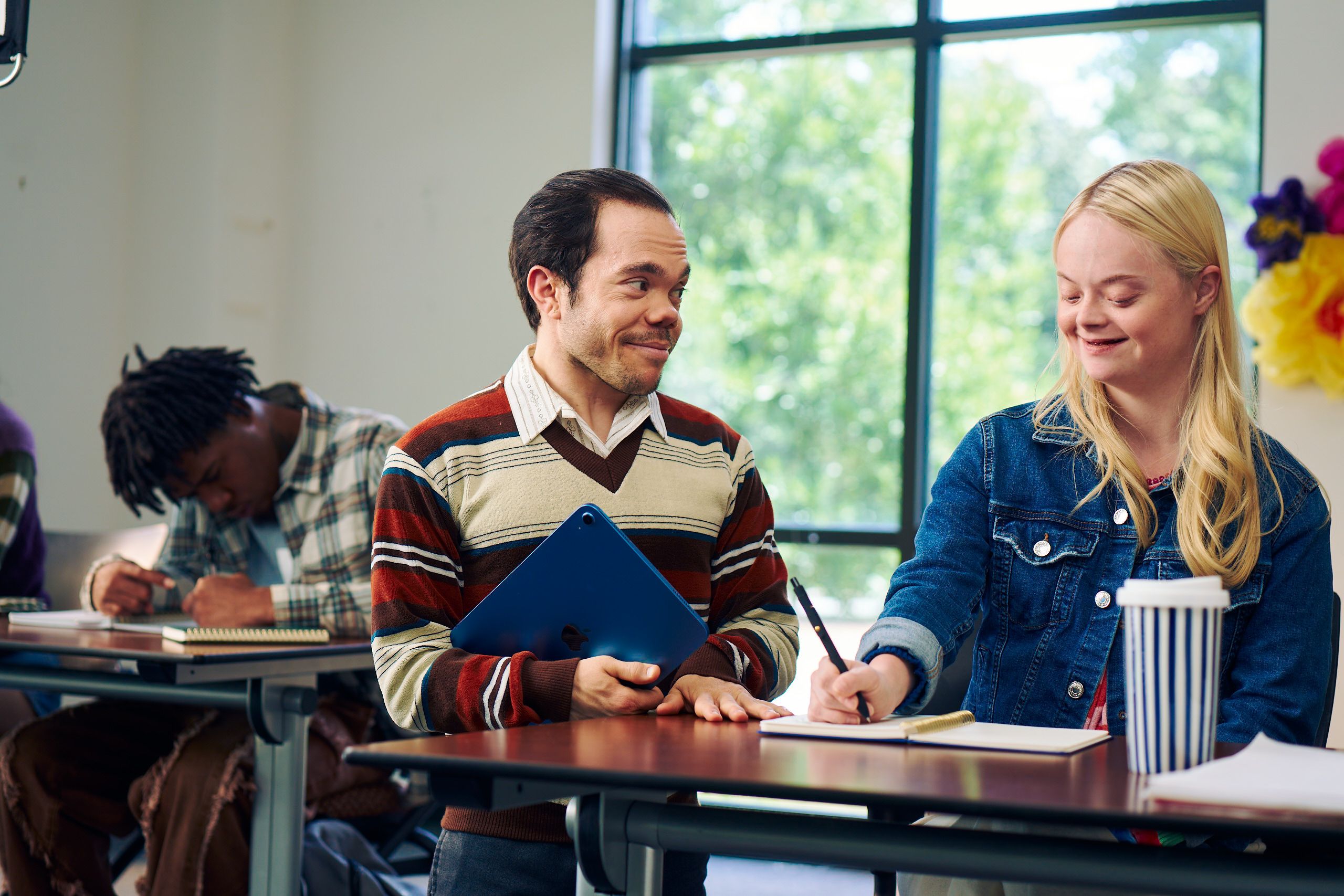 A caucasian college student with Down Syndrome sits at a lecture table and takes notes while her professor, who is standing next to her, chats with her. She has long blonde hair, blue eyes, and is wearing a jean hacket and several bright scrunchies on her wrist. Her professor, a white man with medium brown hair and light brown eyes who has dwarfism, smiles and holds a tablet in his hand. He has short locs and is wearing a red striped sweater, a collared shirt, and dark dress pants. At another table, a Black man with short hair takes notes with focus. A large window with trees showing is at the back of the classroom.