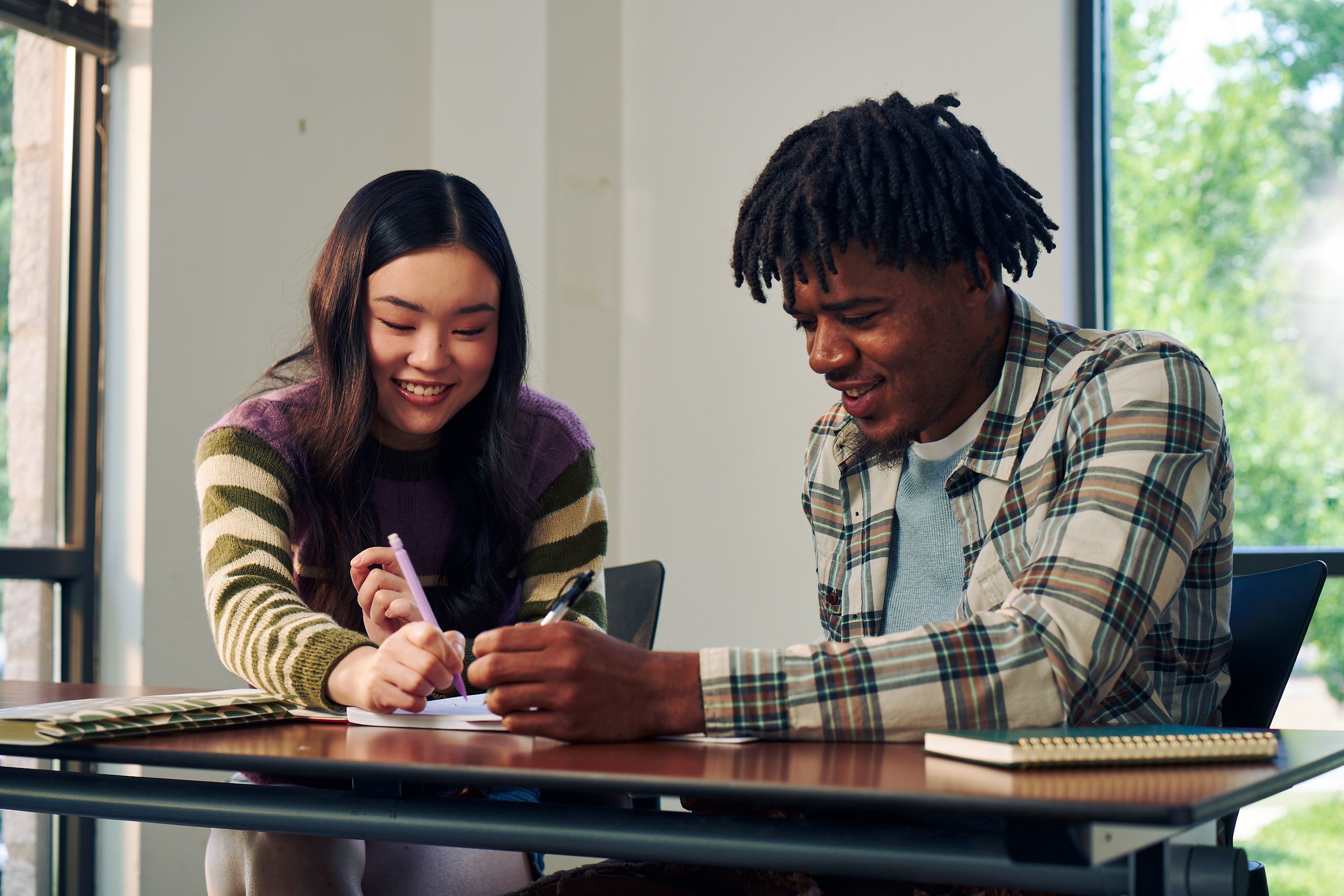 Two college students smile and write in a worksheet together at a lecture table. One student, a Black man, has short locs and is wearing a white plaid shirt and a blue knit shirt underneath. The other student, an East Asian young woman, has long curled hair and is wearing a purple, green, and tan striped sweater.