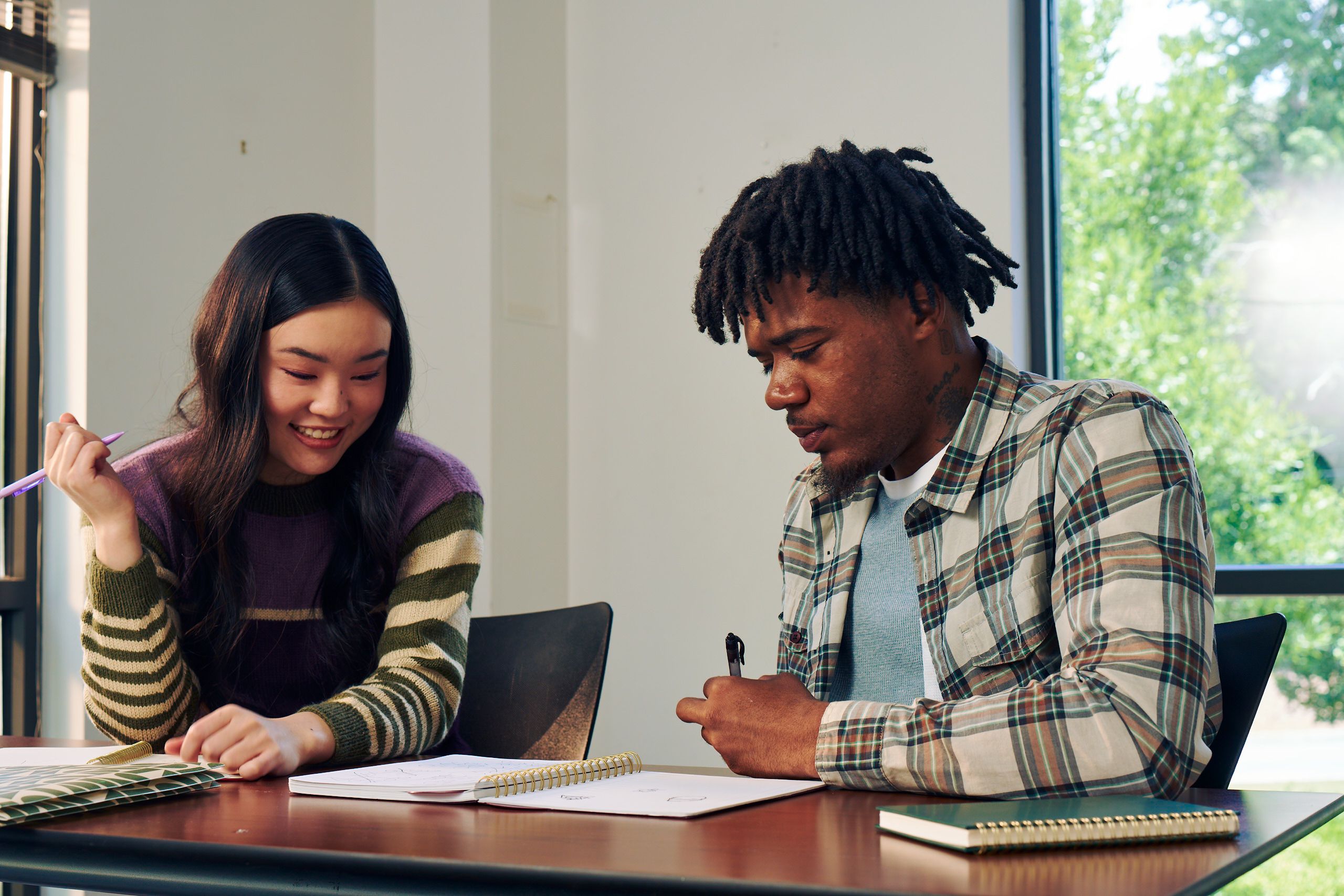 Two college students study notes in an open notebook together at a lecture table. They are both holding pencils. One student, a Black man with short locs is wearing a white plaid shirt and a blue knit shirt underneath. The other student, an East Asian young woman, has long curled hair and is wearing a purple, green, and tan striped sweater.