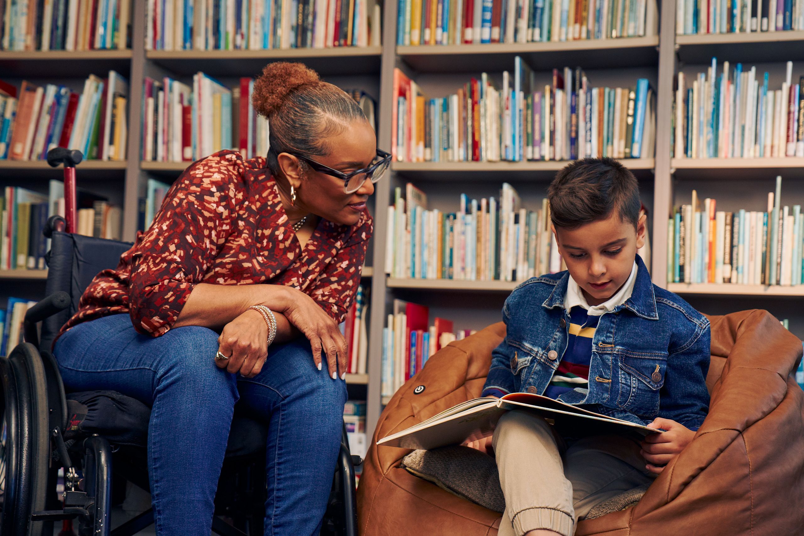 An African American teacher seated in a manual wheelchair watches a young student sitting in a bean bag chair read a picture book. The teacher has medium brown skin, reddish brown hair pulled into a short natural ponytail, and is wearing reading glasses, a red patterned blouse, and jeans. The young student is a Hispanic and Mediterranean-American boy with short brown hair. He has olive skin and is wearing a blue striped shirt, a jean jacket, and khaki pants. A wall of children's books are behind them.