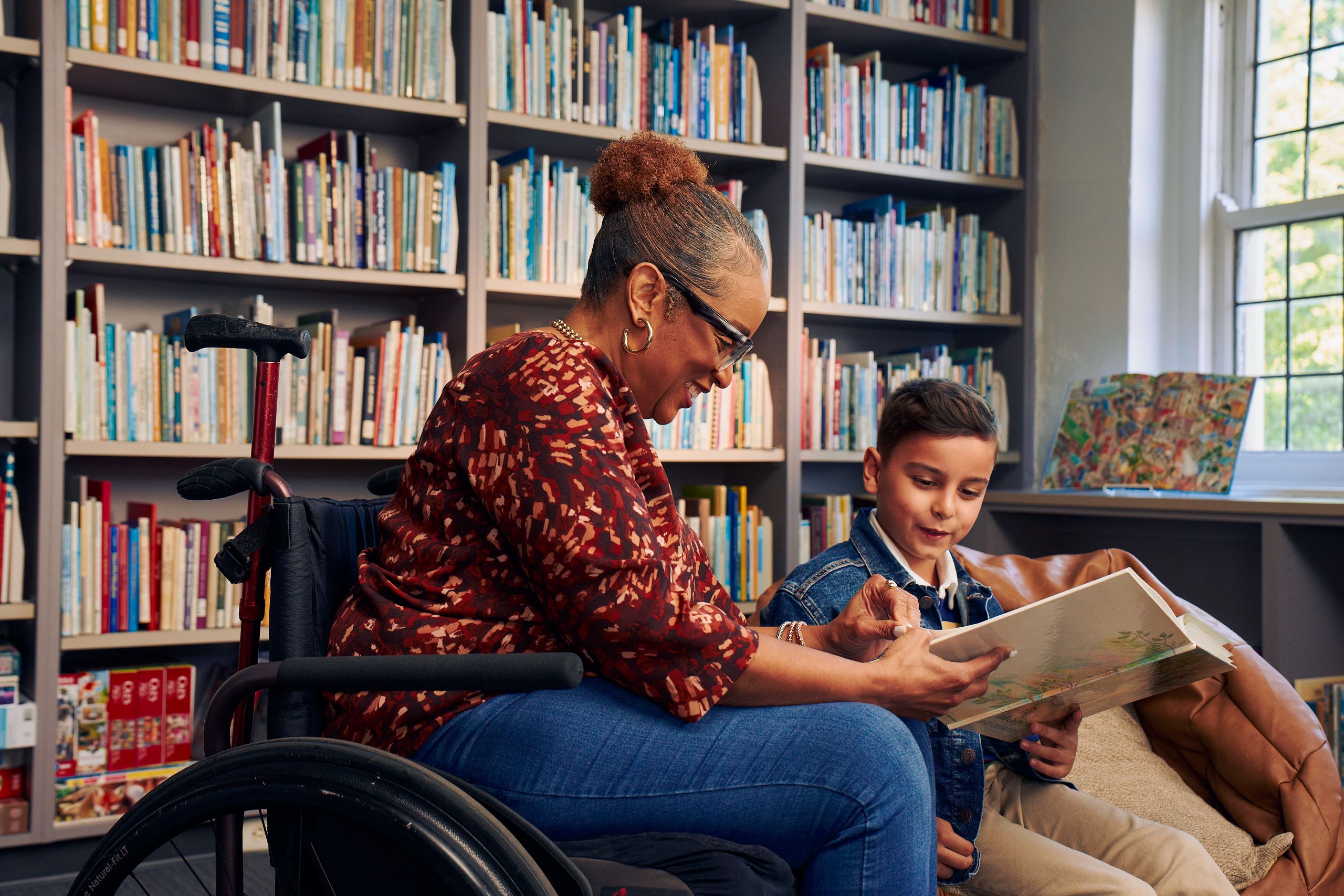 A young boy sitting in a bean bag chair and his African American teacher seated in a manual wheelchair hold and look at a picture book together. The teacher has a red walking cane fastened to the back of her wheelchair. She has medium brown skin, reddish brown hair pulled into a short natural ponytail, and is wearing reading glasses, a red patterned blouse, and jeans. The young student is a Hispanic and Mediterranean-American boy with short brown hair. He has olive skin and is wearing a blue striped shirt, a jean jacket, and khaki pants. A wall of children's books are behind them.