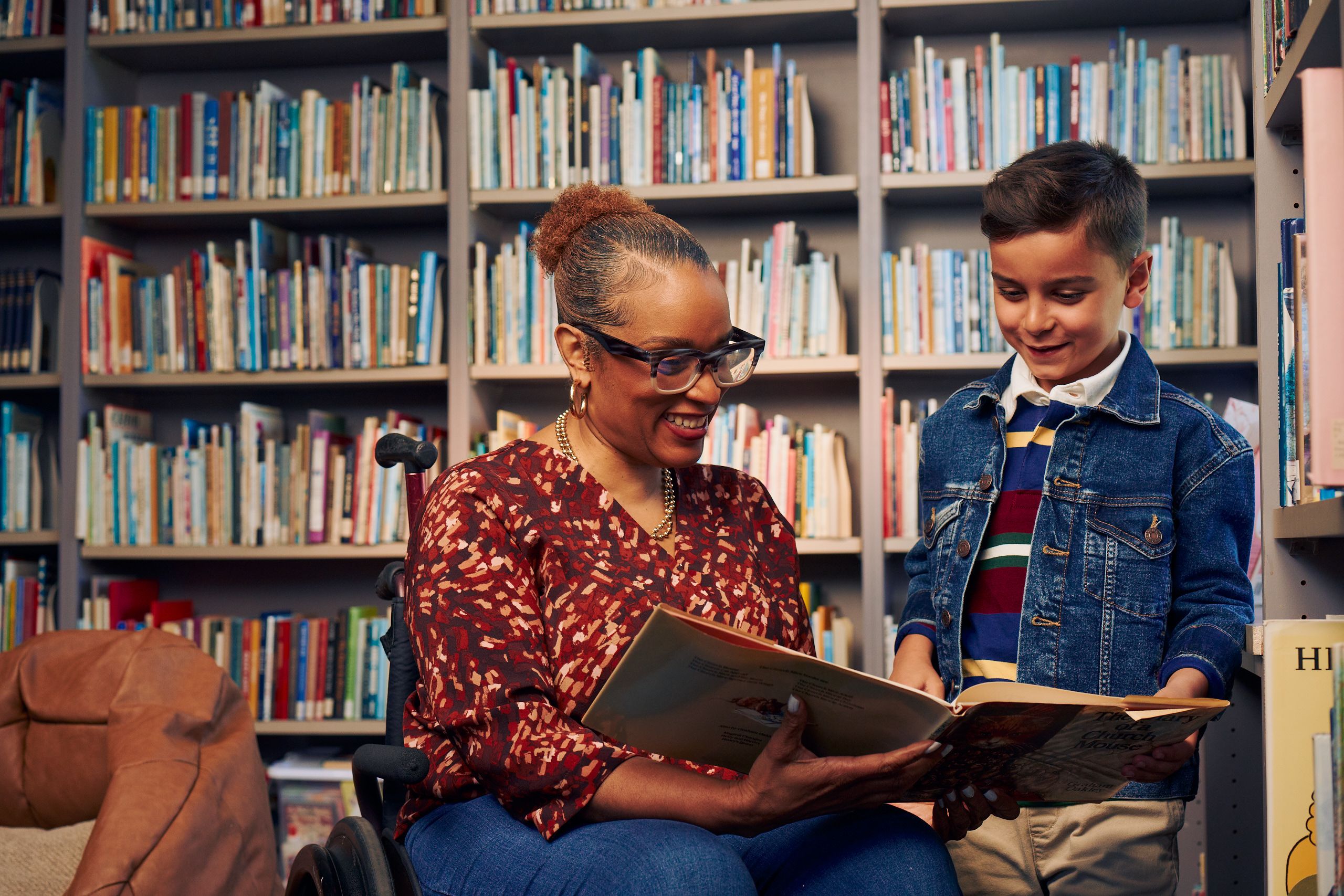 An African American teacher seated in a manual wheelchair hold and her young student and look at a picture book together. The teacher has a red walking cane fastened to the back of her wheelchair. She has medium brown skin, reddish brown hair pulled into a short natural ponytail, and is wearing reading glasses, a red patterned blouse, and jeans. The young student is a Hispanic and Mediterranean-American boy with short brown hair. He has olive skin and is wearing a blue striped shirt, a jean jacket, and khaki pants. A wall of children's books and a brown bean bag chair are behind them.