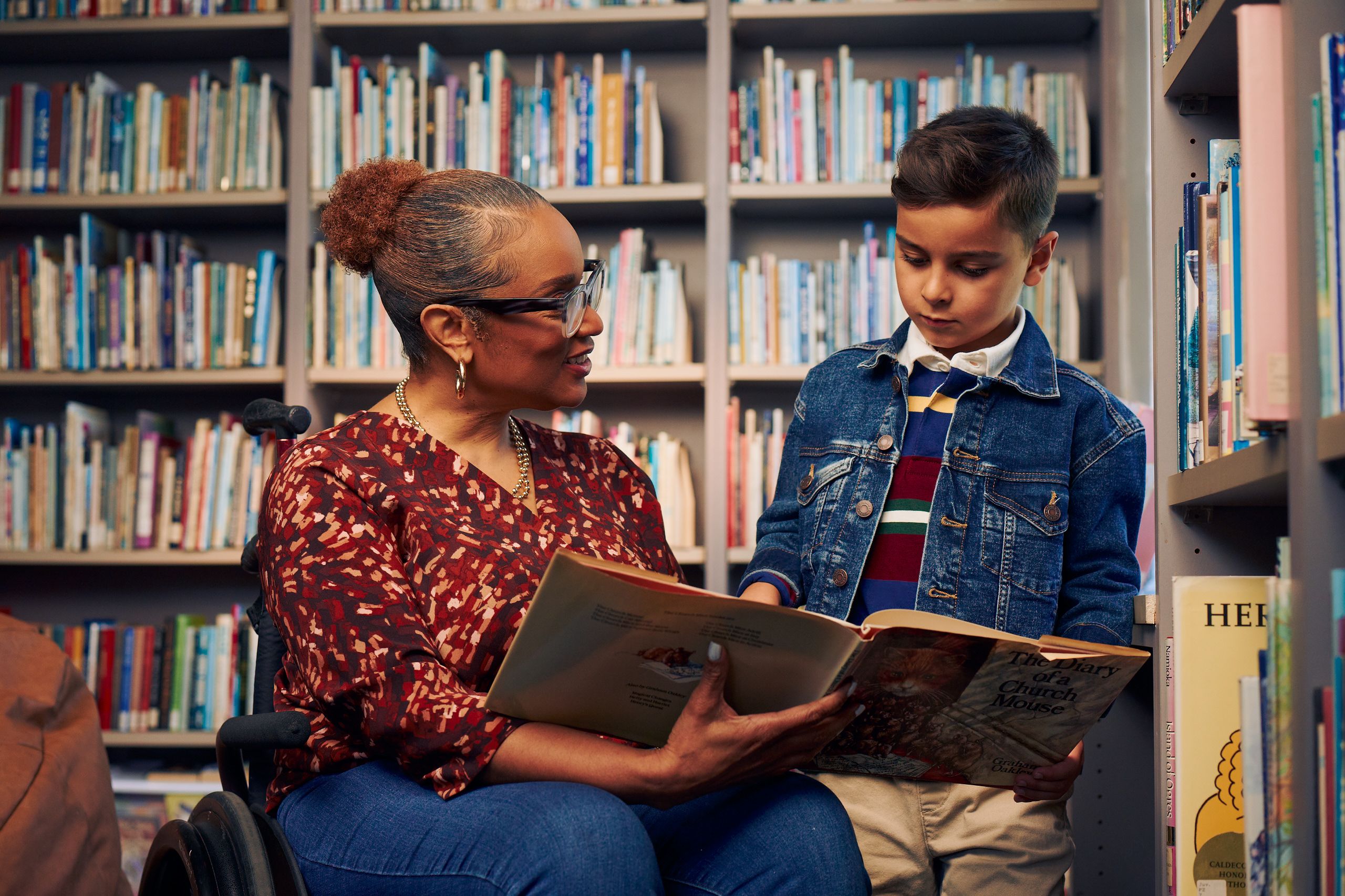 An African American teacher seated in a manual wheelchair holds a picture book open while her young student reads out loud. The teacher has medium brown skin, reddish brown hair pulled into a short natural ponytail, and is wearing reading glasses, a red patterned blouse, and jeans. The young student is a Hispanic and Mediterranean-American boy with short brown hair. He  has olive skin and is wearing a blue striped shirt, a jean jacket, and khaki pants. A wall of children's books are behind them.