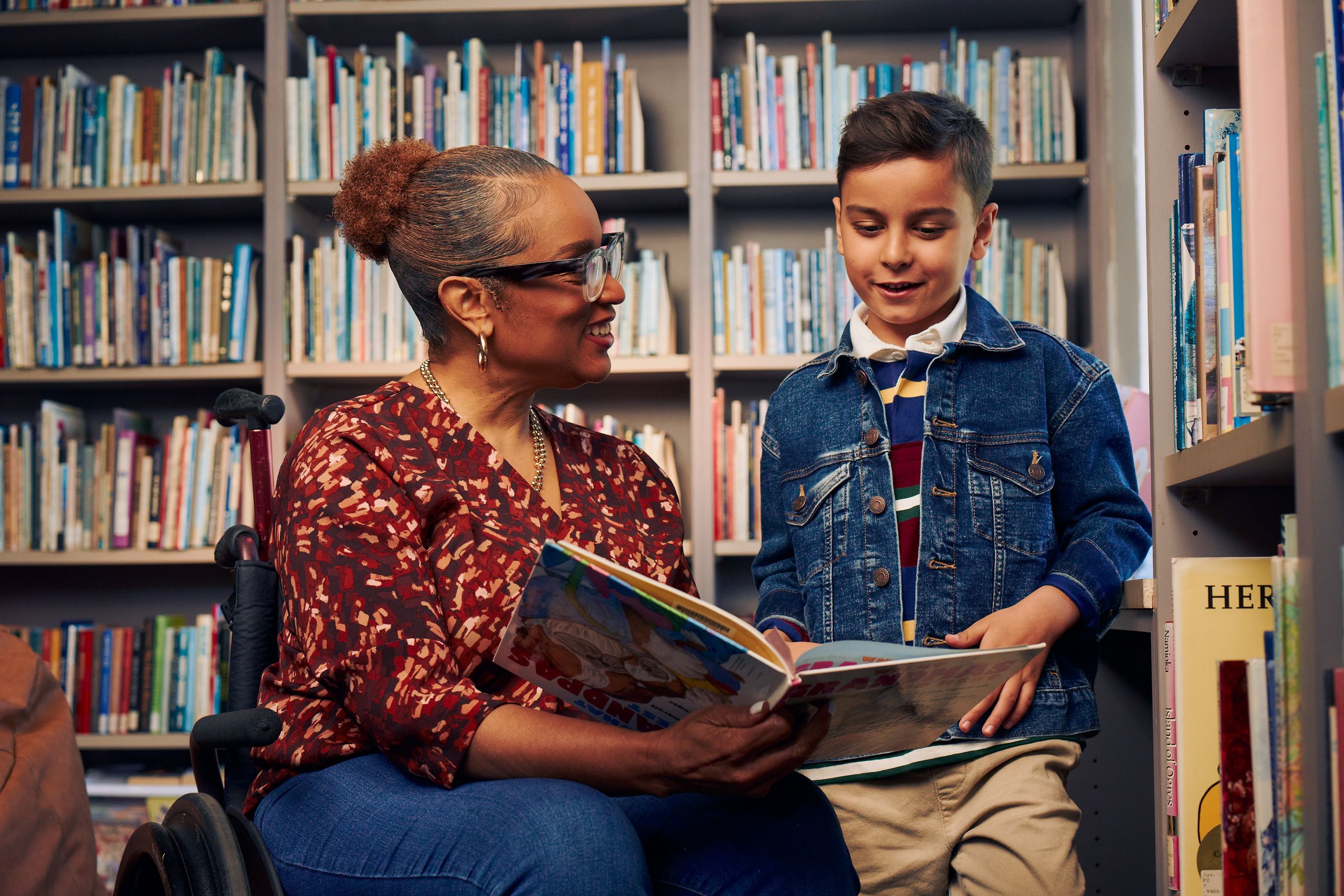 An African American teacher seated in a manual wheelchair smiles and holds a picture book open while her young student reads out loud. The teacher has medium brown skin, reddish brown hair pulled into a short natural ponytail, and is wearing reading glasses, a red patterned blouse, and jeans. The young student is a Hispanic and Mediterranean-American boy with short brown hair. He  has olive skin and is wearing a blue striped shirt, a jean jacket, and khaki pants. A wall of children's books are behind them.