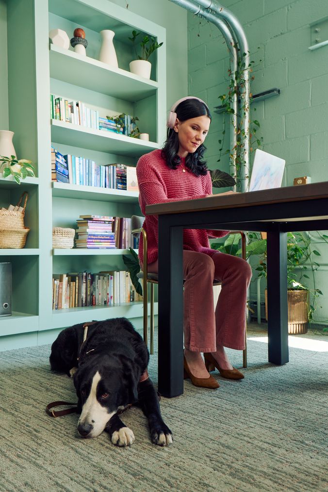 A young pale white blind woman is sitting at a desk typing on a laptop computer in a modern coworking office space. A black and white guide dog wearing a guide harness is lying on the floor near her feet. The woman has shiny black hair styled in waves and is wearing over the ear headphones, a chunky knitted pink sweater, dusky pink flared pants, and taupe pointed toe suede heels. The coworking office space is painted a warm mint green and features a large bookcase with several books, plants, a rattan basket, and clay vases.