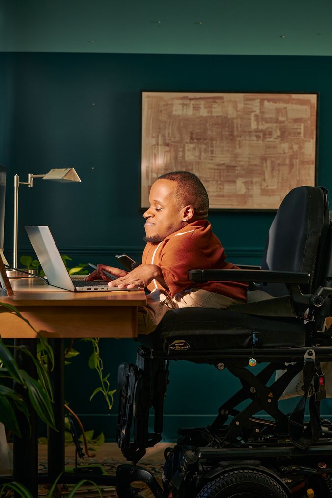 A black man who is a little person is seated in his power wheelchair in a dark teal home office. He types on a laptop on his desk. A lamp and a framed photo are on the desk in front of him. His wheelchair is in an elevated position, allowing him to be level with his desk. His desk is surrounded by several house plants and an abstract art piece. He has closely cropped black hair, brown eyes, and is wearing a rust-orange knit quarter-zip sweater with white vertical stripes, light khaki trousers, and a small stud earring. A tote bag hangs on the back of his wheelchair.