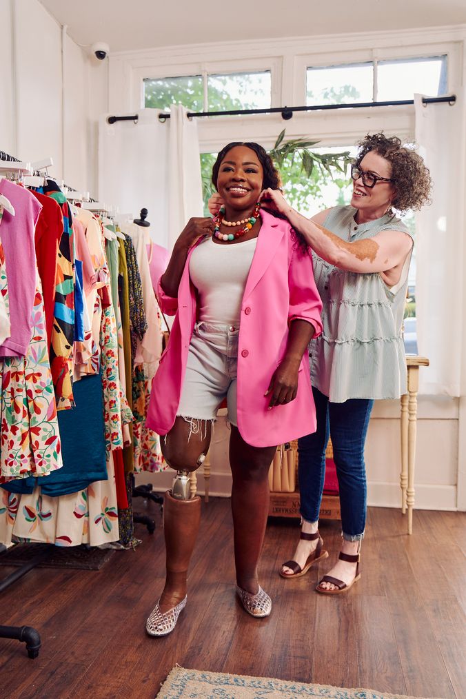 A black woman with a prosthetic leg smiles wide as a Black woman shopkeeper with Vitiligo smiles and helps on a bright necklace in a boutique. The black woman has long, black curly hair and is wearing a vibrant pink blazer, jean cut off shorts, and jelly sandals. The shopkeeper has a short, curly bob and is wearing glasses, a striped flowy blouse, jeans, and brown sandals. A clothing rack filled with high end garments is next to the two women.
