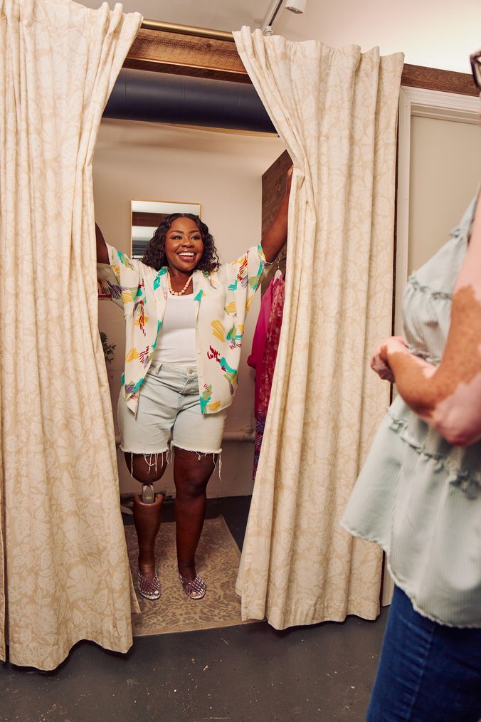 A black woman with a prosthetic leg opens up the curtains of a dressing room of a boutique with a big smile on her face. She is wearing a patterned white blouse, a white tank, jean cut off shorts, and jelly sandals. She looks at the shopkeeper, partially off-camera. Several high end garments are on hooks in the dressing room. The shopkeeper is a Black woman with Vitiligo. She has a short, curly bob and is wearing glasses, a flowy blouse, and jeans.