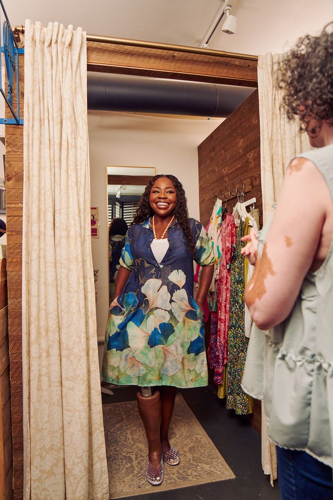A black woman with a prosthetic leg poses in a dressing room of a boutique with a big smile on her face. She is wearing a blue patterned collared dress and jelly sandals. She has her hands in the pockets of the dress and looks at the shopkeeper, partially off-camera. Several high end garments are on hooks in the dressing room. The shopkeeper is a Black woman with Vitiligo. She has a short, curly bob and is wearing glasses, a flowy blouse, and jeans.