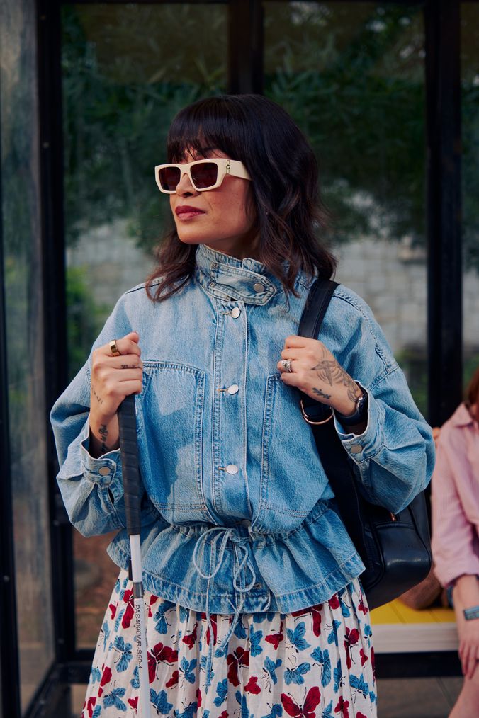 An Asian Filipino blind woman with Retinitis Pigmentosa and Usher Syndrome stands at a covered bus stop. She is wearing white sunglasses and is holding her white cane with her tattooed hands. She has on a jean jacket, a butterfly patterned skirt, and a black backpack.