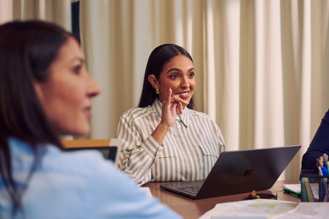 A Deaf Latina Puerto Rican woman uses American Sign Language while seated at a conference room table with two colleagues. She has long deep brown hair and is wearing a striped business shirt and hoop earrings. A laptop and business planning documents are in front of her.