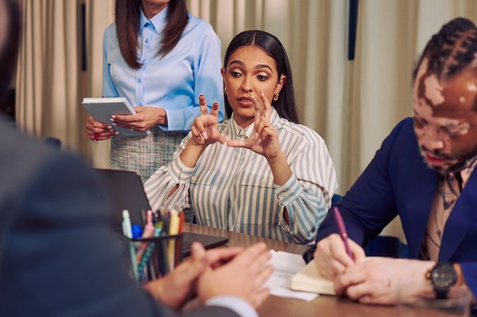 A Deaf Latina Puerto Rican woman uses American Sign Language while seated at a conference room table with three colleagues. She has long deep brown hair and is wearing a striped business shirt and hoop earrings. One colleague, an African American man with olive and porcelain skin, Non-Segmental Vitiligo, and brown barrel-twist dreadlocks writes in a notebook next to her. He is wearing a blue business suit.