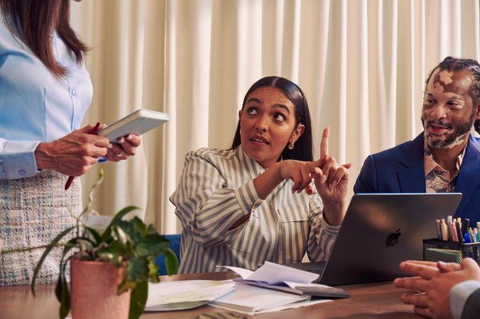 A Deaf Latina Puerto Rican woman communicates in American Sign Language to a Caucasian woman with long straight brown hair wearing business attire standing next to her while seated at a conference room table with two colleagues. The Deaf woman has long deep brown hair and is wearing a striped business shirt and hoop earrings. An African American man with olive and porcelain skin, Non-Segmental Vitiligo, and brown barrel-twist dreadlocks looks at the Deaf woman with focus. A laptop, business planning documents, a cup of pens, and a potted plant are on the conference room table.