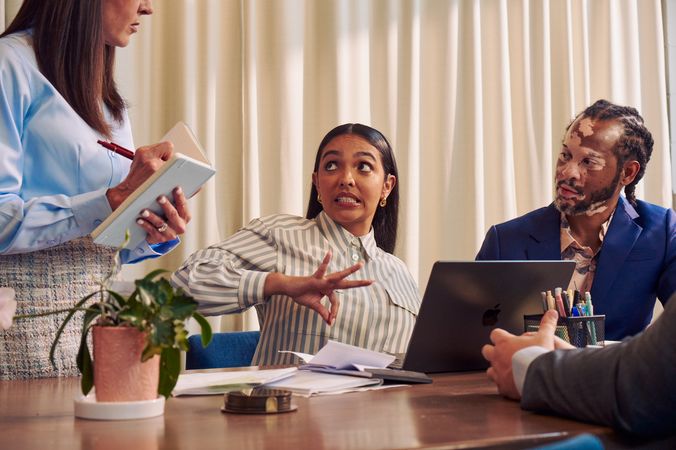A Deaf Latina Puerto Rican woman communicates in American Sign Language to a Caucasian woman with long straight brown hair wearing business attire standing next to her while seated at a conference room table with two colleagues. The Deaf woman has long deep brown hair and wears a striped business shirt and hoop earrings. An African American man with olive and porcelain skin, Non-Segmental Vitiligo, and brown barrel-twist dreadlocks wearing a business suit looks at the Deaf woman with focus. A laptop, business planning documents, a cup of pens, and a potted plant are on the conference room table.