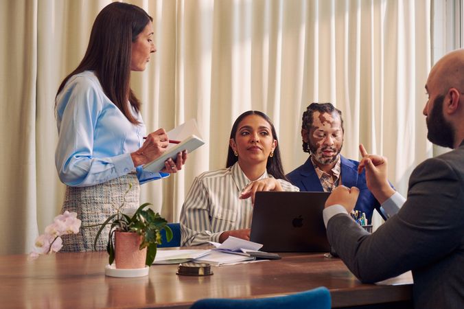 A Caucasian woman with long straight brown hair wearing business attire, holds a notebook and pen while speaking to a Deaf Latina Puerto Rican woman seated next to her. A bald white sign language interpreter sitting across from the women translates the Caucasian woman's speech into American Sign Language, while the Deaf woman looks on. The interpreter is wearing a gray business suit. The Deaf woman has long deep brown hair and is wearing a striped business shirt and hoop earrings. An African American man with olive and porcelain skin, Non-Segmental Vitiligo, and brown barrel-twist dreadlocks wearing a business suit looks downward. A laptop, business planning documents, a cup of pens, and a potted plant are on the conference room table.
