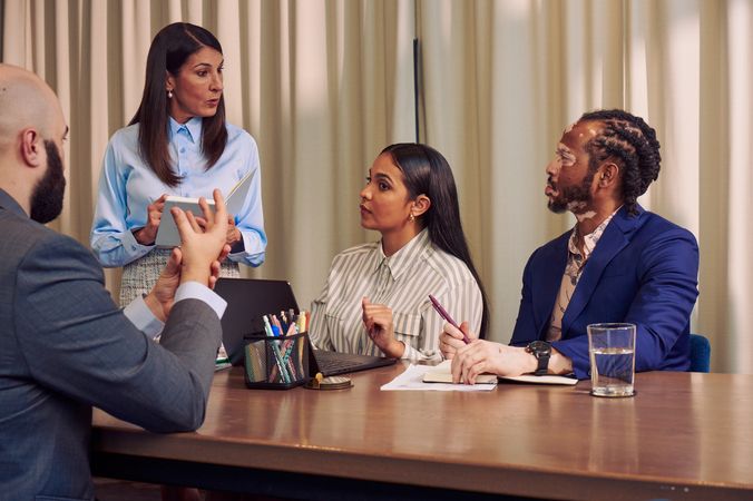 A Caucasian woman with long straight brown hair wearing business attire speaks to a Deaf Latina Puerto Rican woman sitting next to her at a conference room table. The Deaf woman watches a bald white sign language interpreter translate the Caucasian woman's speech into American Sign Language. The Deaf woman has long deep brown hair and is wearing a striped business shirt and hoop earrings. The interpreter is wearing a gray business suit. An African American man with olive and porcelain skin and Non-Segmental Vitiligo looks at the Caucasian woman. The man has brown barrel-twist dreadlocks and is wearing a blue business suit. A laptop, business planning documents, a cup of pens, and a potted plant are on the conference room table.