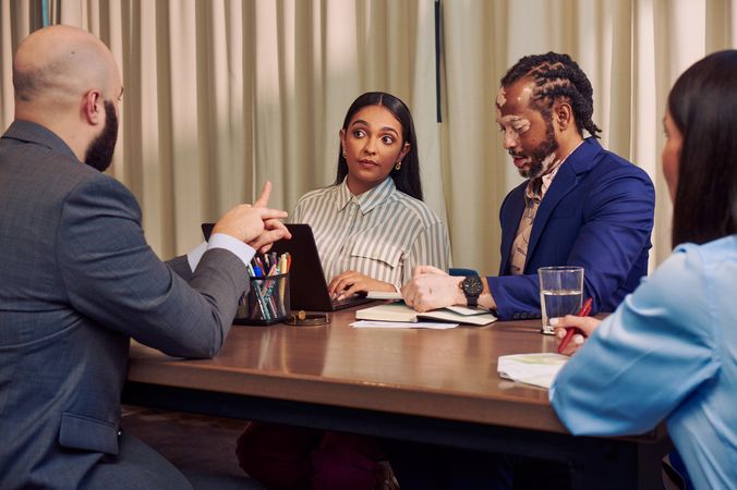 A bald white sign language interpreter translates speech into American Sign Language for a Deaf Latina Puerto Rican woman sitting across from him at a conference room table. An African American man with olive and porcelain skin and Non-Segmental Vitiligo is seated next to her at the table, writing in a notebook. A Caucasian woman with long brown hair writes in a notebook across the table. All four colleagues are wearing business attire.