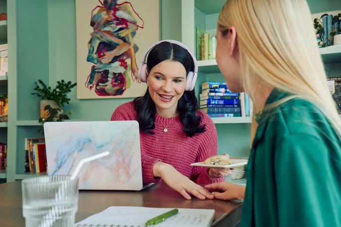 A young pale white blind woman holds a plate with a scone while reaching forward with her other hand towards a white woman with partially paralyzed arms and hands who is sitting next to her at a table in a modern coworking office space. The blind woman has shiny black hair styled in waves and wears a chunky knitted pink sweater and over the ear headphones, which are partially behind one ear. The woman with partially paralyzed arms and hands places her hand over the blind woman's, showing where she is sitting at the table. She is wearing a green blazer. The blind woman has a laptop in front of her. The woman with partially paralyzed arms and hands has a notebook in front of her and a glass of water with a straw in it nearby. The coworking office space is painted a warm mint green and features a large bookcase with several books, plants, and a large abstract art piece.