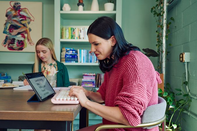 A young pale white blind woman is sitting at a table, typing on a keyboard connected to a tablet computer in a modern coworking office space. Visible on her tablet is a document in large print that reads \"Launch timing: May 11. Rollout plan: Teaser. Drumbeat.\" Across from her at the table, a young white woman with partially paralyzed arms and hands writes in a notebook in front of her. A glass of water with a straw in it is nearby. The woman with partially paralyzed arms and hands has long straight blonde hair and is wearing a floral dress and green blazer. The blind woman has shiny black hair styled in waves and is wearing a chunky knitted pink sweater. The coworking office space is painted a warm mint green and features a large bookcase with several books, plants, clay vases, and a large abstract art piece.
