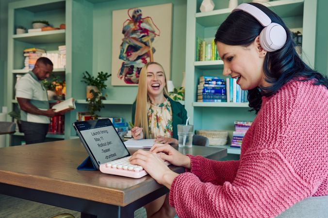 A young pale white blind woman is sitting at a table, typing on a keyboard connected to a tablet computer in a modern coworking office space. Visible on her tablet is a document in large print that reads \"Launch timing: May 11. Rollout plan: Teaser. Drumbeat.\" She is smiling as she types. Across from her at the table, a young white woman with partially paralyzed arms and hands laughs with the blind woman as she holds a pen to a notebook in front of her. A glass of water with a straw in it is nearby. The woman with partially paralyzed arms and hands has long straight blonde hair and is wearing a floral dress and green blazer. The blind woman has shiny black hair styled in waves and is wearing over the ear headphones that partially cover one ear and a chunky knitted pink sweater. The coworking office space is painted a warm mint green and features a large bookcase with several books, plants, and a large abstract art piece.