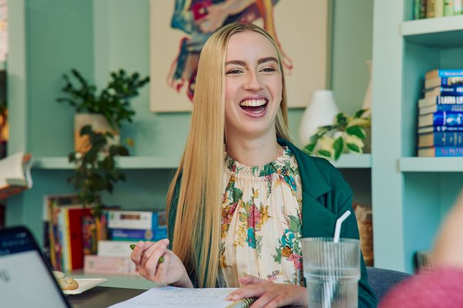 A young white woman with long, straight blonde hair and partially paralyzed arms and hands laughs with a wide smile. She is sitting at a table in a modern coworking office space holding a pen with an open notebook in front of her. A glass of water with a straw in it is nearby. She is wearing a floral dress and a green blazer. The coworking office space is painted a warm mint green and features a large bookcase with several books, plants, and a large abstract art piece.