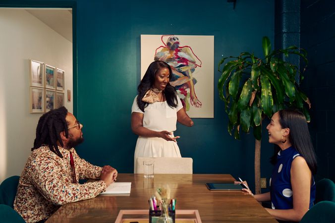 An executive leads a small team meeting in a dark conference room, looking at a Korean and Filipino woman sitting at the conference table in front of her. The executive is a Black American woman and below-elbow congenital amputee wearing a cream blouse and pants, with a patterned scarf around her neck. Her hair is side parted and coiffed. The woman sitting at the table is wearing a blue polka dot blouse sits at the conference table, smiling. Her tablet is in front of her. Across the table sits a neurodivergent Black male with locs with a notebook in front of him. He is wearing glasses, a floral print button down shirt, and a red tie. The conference room is painted dark teal and features an abstract art piece and a large potted plant. The conference room door is open to a long white hallway with framed art pieces hanging.