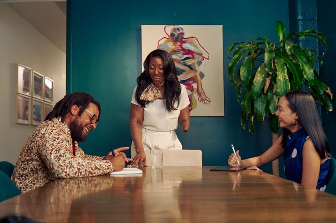 An executive leads a small team meeting in a dark conference room, smiling and looking down at a notebook that a neurodivergent Black male seated at the conference table is writing in. The executive is a Black American woman and below-elbow congenital amputee wearing a cream blouse and pants, with a patterned scarf around her neck. Her hair is side parted and coiffed. The Black male has locs and is wearing glasses, a floral print button down shirt, and a red tie. Across the table sits a Korean and Filipino woman wearing a blue polka-dot blouse, smiling. Her tablet is in front of her. The conference room is painted dark teal and features an abstract art piece and a large potted plant. The conference room door is open to a long white hallway with framed art pieces hanging.