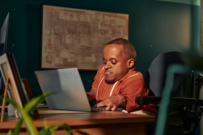 A black man who is a little person is seated in his power wheelchair in a dark teal home office. He types on a laptop on his desk, with a monitor and framed photos nearby.  An abstract art piece is on the wall behind him. He has closely cropped black hair, brown eyes, and is wearing a rust-orange knit quarter-zip sweater with white vertical stripes, light khaki trousers, and a small stud earring.