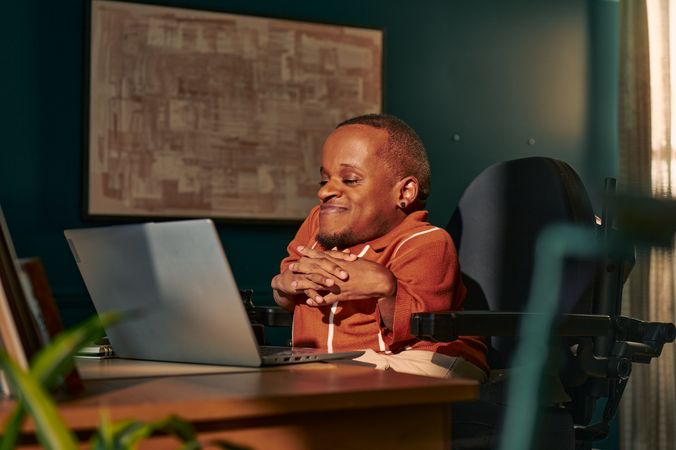 A black man who is a little person is seated in his power wheelchair in a dark teal home office. He smiles down at his open laptop, his hands together with fingers interlaced. A  notebook and framed photos are on the desk in front of him. An abstract art piece is on the wall behind him. He has closely cropped black hair, brown eyes, and is wearing a rust-orange knit quarter-zip sweater with white vertical stripes, light khaki trousers, and a small stud earring.