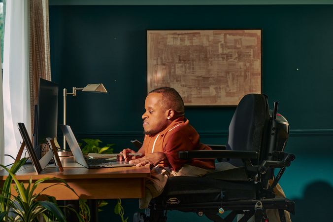 A black man who is a little person is seated in his power wheelchair in a dark teal home office. He holds a pen and types on his laptop. A monitor, notebook, pen, lamp, and framed photos are on the desk in front of him. His wheelchair is in an elevated position, allowing him to be level with his desk. He is facing a window, letting in light through white and patterned curtains. His desk is surrounded by several house plants and an abstract art piece. He has closely cropped black hair, brown eyes, and is wearing a rust-orange knit quarter-zip sweater with white vertical stripes, light khaki trousers, and a small stud earring. A tote bag hangs on the back of his wheelchair.