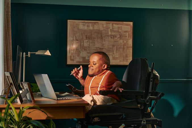A black man who is a little person is seated in his power wheelchair in a dark teal home office. He holds a pen and looks down at his laptop. A monitor, notebook, pen, lamp, and framed photos are on the desk in front of him. His desk is surrounded by house plants and an abstract art piece. He has closely cropped black hair, brown eyes, and is wearing a rust-orange knit quarter-zip sweater with white vertical stripes, light khaki trousers, and a small stud earring. A tote bag hangs on the back of his wheelchair.