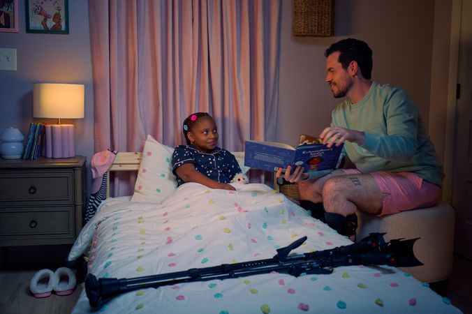 A white man with cerebral palsy who wears leg braces holds a children's book titled "Extraordinary Jordyn and Her Bionic Ears" while sitting on an ottoman stool in his daughter's bedroom at night. His daughter is a Black multiracial American girl with thick braided red hair adorned with two flower barrettes. She is tucked into bed and is wearing dark blue patterned pajamas and looks up at her father with a soft smile. The father's black forearm crutches lie on the foot of the bed. He has dark brown curly hair, short brown facial hair, and is wearing a light green sweater with pink shorts. He has a tattoo of the Roman numeral two followed by a slash and the number three. The child's room is painted light pink and includes a nightstand, lamp, a pair of slippers, curtains, and children's books. Her bedspread is white with blue, pink, and yellow polka dots. Framed illustrations of Black women hang on the wall.