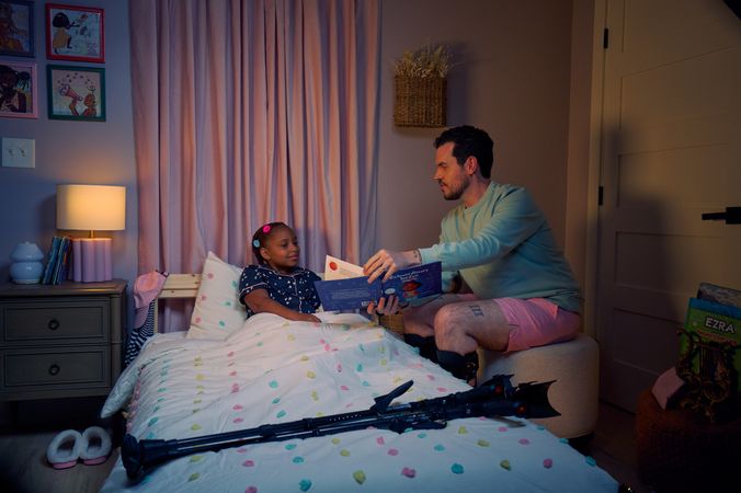 A white man with cerebral palsy who wears leg braces shows a children's book titled "Extraordinary Jordyn and Her Bionic Ears" to his daughter in her bedroom at night. His daughter is a Black multiracial American girl with thick braided red hair adorned with two flower barrettes. She is tucked into bed and is wearing dark blue patterned pajamas and looks at the open book. The father's black forearm crutches lie on the foot of the bed. He has dark brown curly hair, short brown facial hair, and is wearing a light green sweater with pink shorts. He has a tattoo of the Roman numeral two followed by a slash and the number three. The child's room is painted light pink and includes a nightstand, lamp, a pair of slippers, curtains, and children's books. Her bedspread is white with blue, pink, and yellow polka dots. Framed illustrations of Black women hang on the wall.