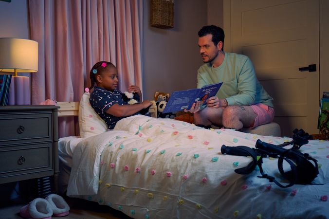 A white man with cerebral palsy who wears leg braces holds a children's book titled "Extraordinary Jordyn and Her Bionic Ears" to his daughter in her bedroom at night. His daughter is a Black multiracial American girl with thick braided red hair adorned with two flower barrettes and is wearing blue patterned pajamas. She is tucked into bed, holding a plushie animal and looks at the open book. The father's black forearm crutches lie on the foot of the bed. He has dark brown curly hair, short brown facial hair, and is wearing a light green sweater with pink shorts. He has a tattoo of the Roman numeral two followed by a slash and the number three. The child's room is painted light pink and includes a nightstand, lamp, a pair of slippers, curtains, and children's books. Her bedspread is white with blue, pink, and yellow polka dots. Framed illustrations of Black women hang on the wall.