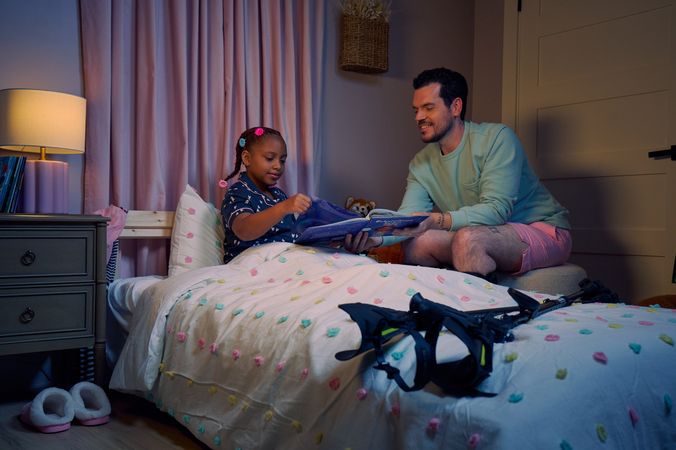 A white man with cerebral palsy who wears leg braces holds a children's book titled "Extraordinary Jordyn and Her Bionic Ears" to his daughter in her bedroom at night. His daughter is a Black multiracial American girl with thick braided red hair adorned with two flower barrettes and is wearing blue patterned pajamas. She is tucked into bed, holding a plushie animal and turns a page in the book. The father's black forearm crutches lie on the foot of the bed. He has dark brown curly hair, short brown facial hair, and is wearing a light green sweater with pink shorts. He has a tattoo of the Roman numeral two followed by a slash and the number three. The child's room is painted light pink and includes a nightstand, lamp, a pair of slippers, curtains, and children's books. Her bedspread is white with blue, pink, and yellow polka dots. Framed illustrations of Black women hang on the wall.