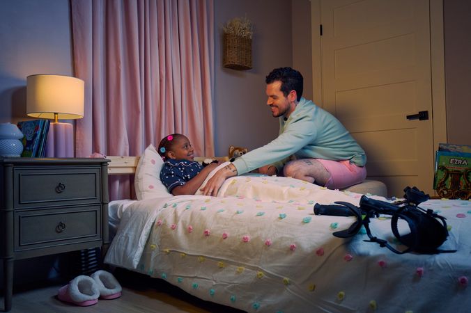A white man with cerebral palsy who wears leg braces tucks his daughter into bed. His daughter is a Black multiracial American girl with thick braided red hair adorned with two flower barrettes and is wearing blue patterned pajamas. She is smiling in bed, and has several plushie animals nearby. The father's black forearm crutches lie on the foot of the bed. He has dark brown curly hair, short brown facial hair, and is wearing a light green sweater with pink shorts. He has a tattoo of the Roman numeral two followed by a slash and the number three. The child's room is painted light pink and includes a nightstand, lamp, a pair of slippers, curtains, and a stack of children's books. Her bedspread is white with blue, pink, and yellow polka dots.