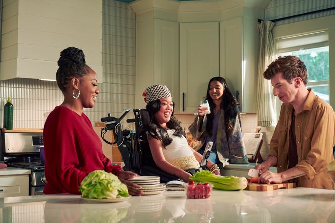 A Southeast Asian woman with curled black hair seated in her power wheelchair smiles and watches her roommate chop vegetables in their bright kitchen. Her smart phone is mounted to her wheelchair. Her roommate is a white male with Moebius Syndrome and a face that is paralyzed. The Southeast Asian woman is wearing a white top with striped, yellow linen pants and a checkered headscarf. He has short, wavy, reddish brown hair and is wearing a tan button down shirt with the sleeves rolled up, showing arm tattoos. Two other roommates laugh and hang out nearby. One roommate has long dark hair and is wearing a purple floral dress, the other is a black female with a braided bun and maroon top.
