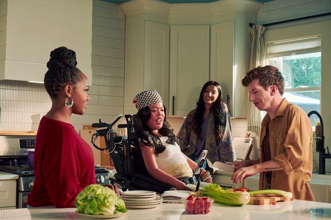 Four roommates hang out in their bright kitchen. One roommate, a white male with Moebius Syndrome and a face that is paralyzed, chops vegetables. Another roomate, a Southeast Asian woman with curled black hair seated in her power wheelchair, watches intently. A third roommate with dark hair wearing a purple floral dress sits on a counter and smiles while watching him prepare the vegetables. A fourth roommate, a black female with a braided bun wearing a maroon top, chats with him.