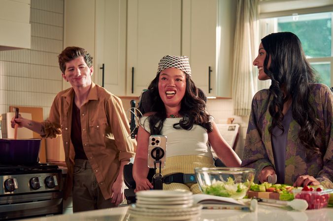 A Southeast Asian woman with curled black hair seated in her power wheelchair smiles brightly in her bright kitchen. Her two roommates are prepping a meal with her. One roommate, a white male with Moebius Syndrome and a face that is paralyzed, smiles at her and stirs a pot on the stove. The other roommate chops vegetables for a salad. The Southeast Asian woman is wearing a white top with striped, yellow linen pants and a checkered headscarf. He has short, wavy, reddish brown hair and is wearing a tan button down shirt with the sleeves rolled up, showing arm tattoos. The third roommate has long dark hair and is wearing a purple floral dress.