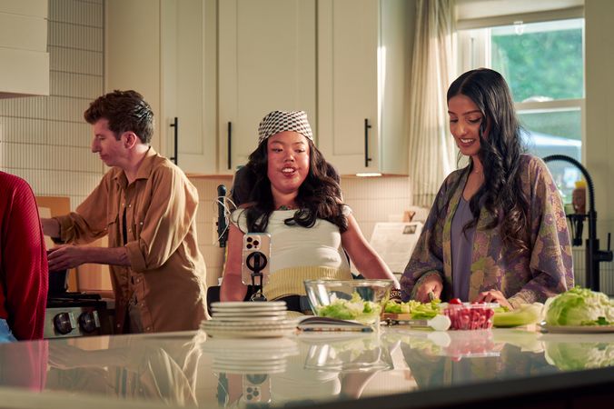 A Southeast Asian woman with curled black hair seated in her power wheelchair watches her roommate prepare a salad in their bright kitchen. Her smart phone is mounted to her wheelchair. Behind them, a white male with Moebius Syndrome stirs a pot on the stove. The Southeast Asian woman is wearing a white top with striped, yellow linen pants and a checkered headscarf. Her roommate has long dark hair and is wearing a purple floral dress.