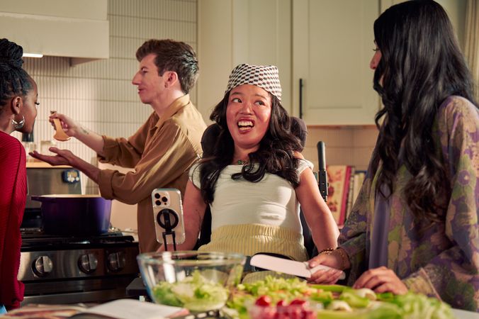 A Southeast Asian woman with curled black hair seated in her power wheelchair smiles widely at her roommate, who is preparing a salad in their bright kitchen. Her smart phone is mounted to her wheelchair. Behind them, a white male with Moebius Syndrome and a face that is paralyzed offers a spoon with sauce to another roommate to taste. The Southeast Asian woman is wearing a white top with striped, yellow linen pants and a checkered headscarf. Her roommate has long dark hair and is wearing a purple floral dress.