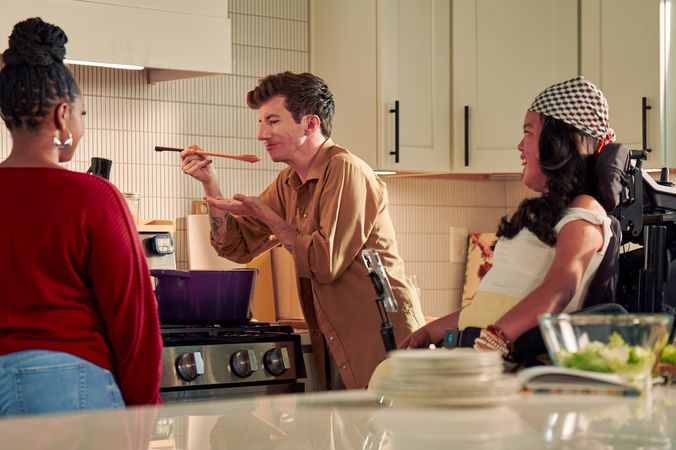 A white male with Moebius Syndrome and a face that is paralyzed cooks in a bright kitchen with his two roommates. He is standing over a stove with a purple pot, holding a spoon to his mouth, about to taste a sauce. Both roommates face him, watching the taste test. One roommate is a tan Southeast Asian woman seated in her power wheelchair; the other is a black female. The male has short, wavy, reddish-brown hair and blue eyes. He is wearing a tan button-down shirt with the sleeves rolled up, showing arm tattoos. The Southeast Asian woman has curled, black hair and is wearing a white top with striped, yellow linen pants, and a checkered head scarf. Her smart phone is mounted to her wheelchair.