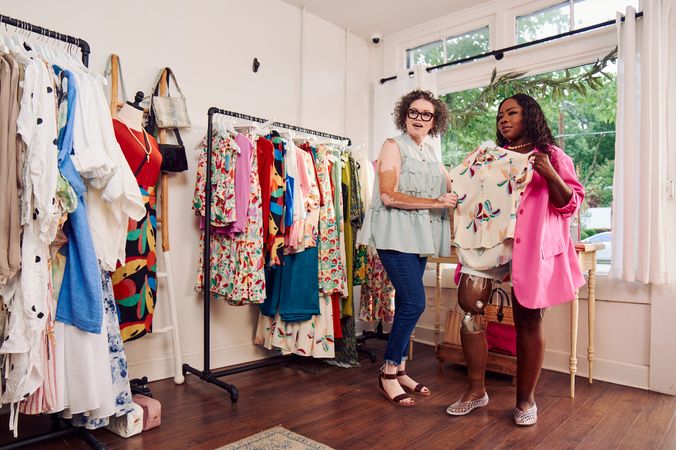 A black woman with a prosthetic leg holds up a patterned blouse to her chest, studying how it would look on her. A Black woman shopkeeper with Vitiligo smiles and helps her hold the garment. The black woman has long, black curly hair and is wearing a vibrant pink blazer, jean cut off shorts, and jelly sandals. The shopkeeper has a short, curly bob and is wearing glasses, a striped flowy blouse, jeans, and brown sandals.Two clothing racks filled with high end garments are next to the two women.